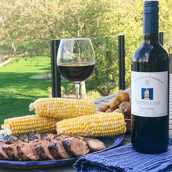 Michele Chiarlo Barbera d'Asti "Le Orme" red wine bottle from Piedmont, Italy standing next to a glass of red wine, corn on the cob and grilled beef
